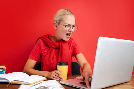 overworked unhappy and frustrated young caucasian woman in stress sitting at office desk, she has a lot of tasks in work, deadline, isolated red backgroundの写真素材