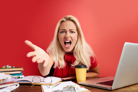 disappointed caucasian woman is dazed and confused by a mistake in official documents at work, sit at desk with laptop and papers, isolated over red backgroundの写真素材