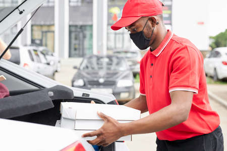 confident deliveryman in red uniform puts orders in the car, he is going to carry it to clients. during quarantineの写真素材
