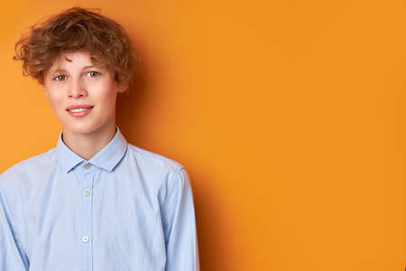 portrait of calm caucasian boy looking at camera, male teenager in blue shirt isolated over orange backgroundの写真素材