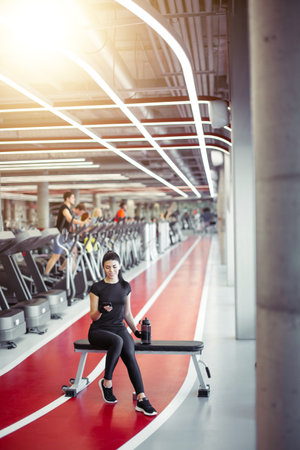 Fit young woman resting after hard training, drink water and looking at smartphone with exercise app