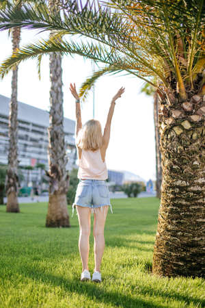 Young female runner in summer active clothes stretching arms before running at the sunny morning standing near big palm in hotel gardenの写真素材