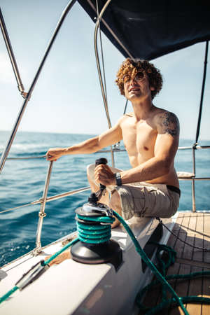Young handsome hipster sailor man with fuzzy hairs sailing a boat to take his friends to a sea trip, looking at horizon, pulling the rope by a winchの写真素材
