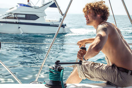 Young handsome hipster sailor man with fuzzy hairs sailing a boat to take his friends to a sea trip, looking at horizonの写真素材