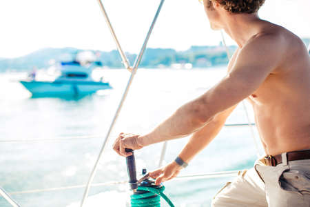 Curly sailor man with naked torso manages with tackle of private motor boat on blue calm water Mediterranean seaの写真素材