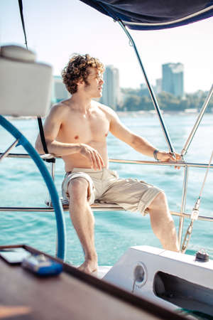 Young red-head hipster man with frizzy hair sitting without shirt, relaxing happily on the vacation sailboat yacht having a rest on summer boat over blue ocean wave backgroundの写真素材