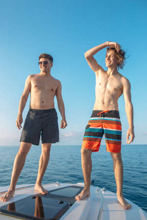 Two happy caucasian male friends traveling on a yacht at open sea, sunbathing on the bow of the boat. Leisure and fun on sea.の写真素材