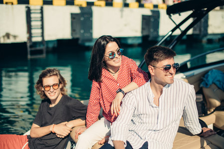 Portrait of happy young fashion woman sitting with two male friends on the motorboat, anchored to a...