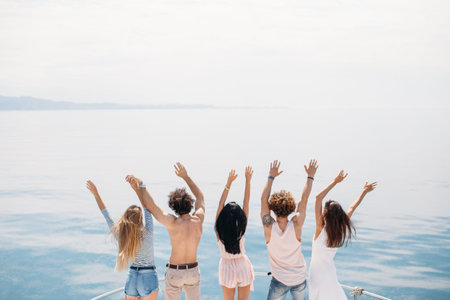 Group of caucasian guys and their girlfriends standing with raised hands on the elge of yacht s...