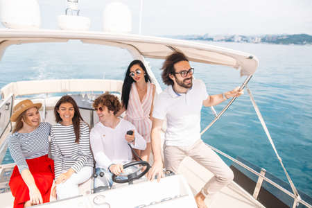 Young curly man navigating a seaboat, sailing in company of his joyful friends, guys and girls sitting beside, enjoying fresh sea windの写真素材