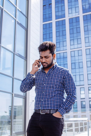 Indian serious man in casual cloth with beard calling with his phone near his ear standing in front...