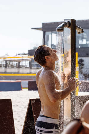 Handsome young topless man with muscular body taking an outdoor shower after sea water over sandy court background. Health and hygiene concept.の写真素材