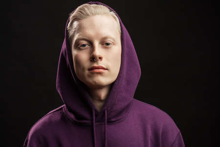 Headshot of young blonde male in purple sportwear with hood over head looking at camera with an expression of superiority isolated on black background.の写真素材