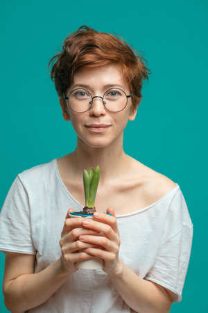 attractive woman with short ginger haircut isolated over blue background with copyspace. Beautiful trendy hipster-looking young female holding small flower in pot.の写真素材
