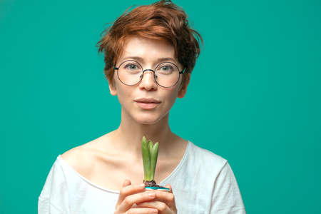Hipster girl with glasses and short red hair dressed in a white T-shirt, standing on the blue background with poted plant in handsの写真素材