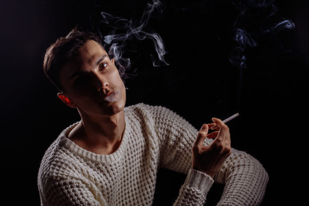 Thougtful young man, wearing knitted white sweater smoking sigarette, thinking, looking at camera on...