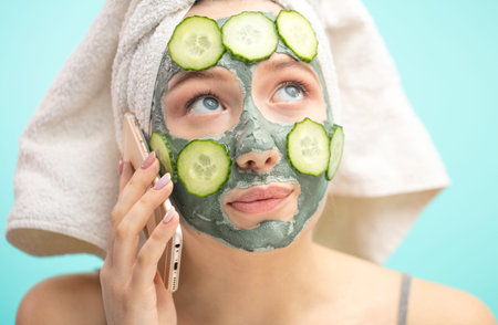 Caucasian woman with towel on head and face mask enriching with cucumbers preparing for a date,...