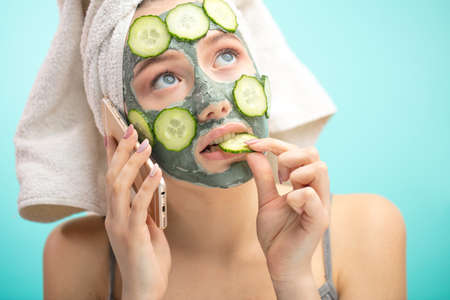 Young healthy caucasian woman with towel on head, making treatments and face clay mask with organic ingredients, chatting on smartphone and eating a slice of cucumber with a pensive expressionの写真素材
