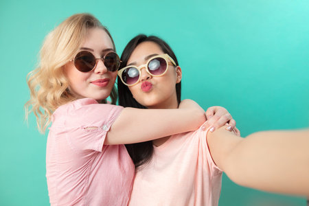 Portrait of pretty charming girls in casual outfits, wearing sunglasses shooting selfie , making...