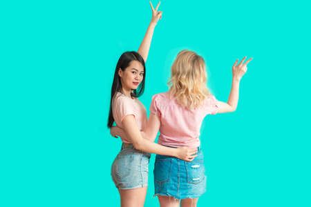 back studio shot of two young mix-race girlfriends, pointing with raised arms to blank space on blue background. Asian woman turns back, looking at cameraの写真素材