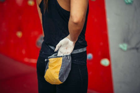 Rear view of unrecognizable female applying magnesium chalk powder on hands from a bag tied to her waist, close up over red and white climbing wall background.の写真素材