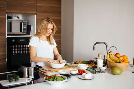 Portrait of happy female wife in white t-shirt cooking healthy dinner at cozy kitchen after workの写真素材