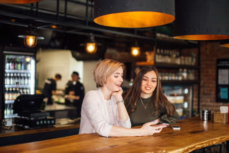 Two attractive excited young using smartphone while sitting at the cafe with cosy stylish interiorの写真素材