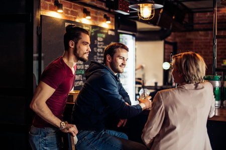 Rear view of two young gyus meeting with a blonde woman in a loft cafe. Man and woman sitting at the...