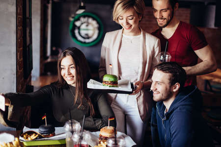 Group of positive cheerful friends having fun, taking selfie with smartphone in burger bar. Young blonde woman holding tray with ready green burgerの写真素材