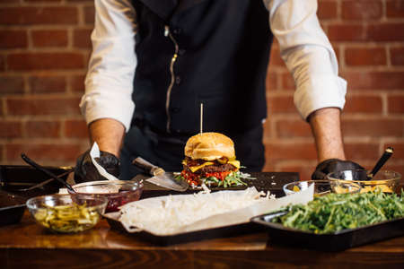 Process of cooking burgers. Cropeed view of chef hands in black gloves with prepared cheeseburger, variety of fillings and ingridients on wooden desk. Catering and Good Quality Fastfood conceptの写真素材
