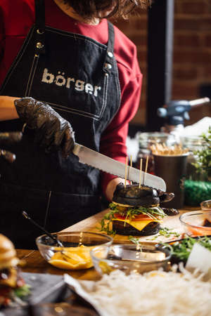 Cropped view of Chef cutting with a knife freshly made black burger with beef chop, salad and vegetables.の写真素材