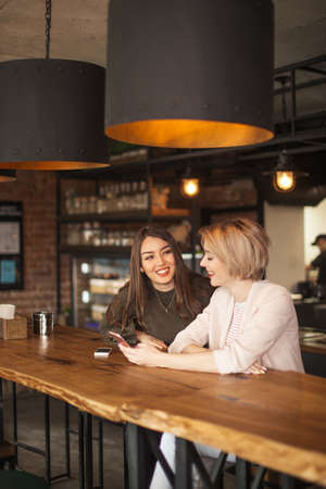 Two cheerful young female students sitting at the Burger snack bar after daytime lections. One of them blogging in social mediaの写真素材
