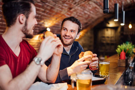 Two happy male friends having dinner at popular burger bar. Young people sitting, talking and eating burgers, drinking beer.の写真素材