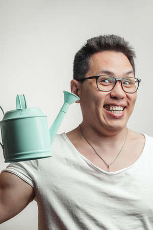 Happy funny Korean man in white common t-shirt and eye glasses with small watering can standing against a white background, looking at camera.の写真素材