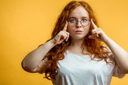 Young attractive woman in spectacles standing thinking with her finger raised totemples and a grimace of concentration as if trying to rememeber something over yellow background with copyspace.の写真素材