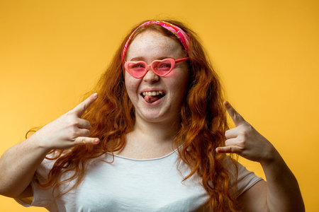 Closeup portrait of a beautiful smiling hipster redhead girl wearing big pink glasses, sticking tongue and gesturingの写真素材