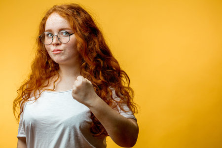 Serious angry woman puts up fist as tries to warn you, isolated over yellow background. Pleasant...