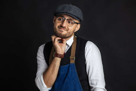 Portrait of pondering young elegant craftsman in classy wear and stylish cap, wearing apron, having doubting or pensive expression, looking away against black backgroundの写真素材