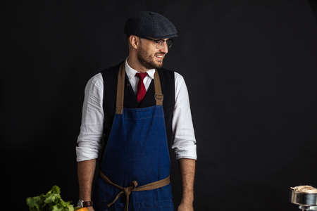 Smiling charismatic male Cook in creative cap and blue apron stands against black wall near a wooden table with ingredients for burgers.の写真素材