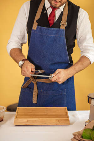 Restaurant Director in classy clothes wearing blue apron, performing masterclass with kitchen utensils knife and sharpen steel tool isolated over yellow backgroundの写真素材