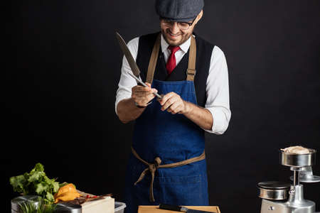 Portrait of chef cook in elegant uniform wearing cap and apron sharpens knife at the restaurant kitchen table over black background. Small business, Service and Catering Conceptの写真素材