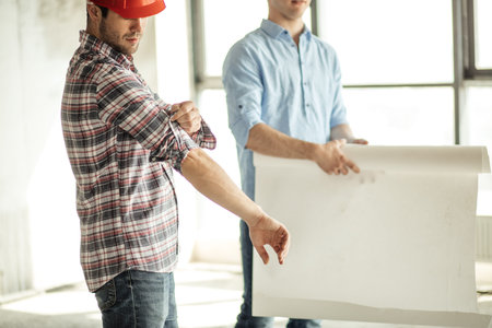 hardworking builder is rolling up his sleevers while his friend is holding a plan of work. close up...