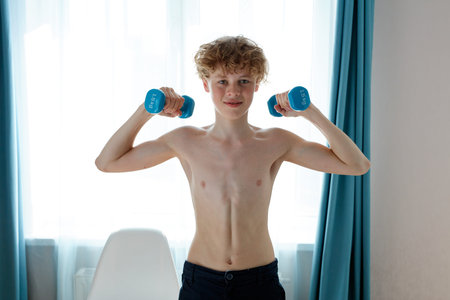 shirtless young caucasian strong teenager boy exercising at home, hold dumbbells in hands, pumping...