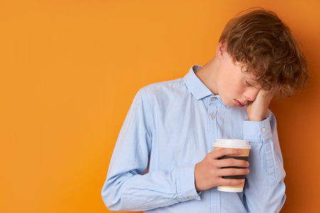 portrait of sleepy teenager boy with cup of coffee isolated on orange background, want to sleep