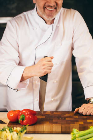 closeup cropped shot of smiling old cook holding the knife on the cutting board indoors. stick a knife.force a knifeの写真素材