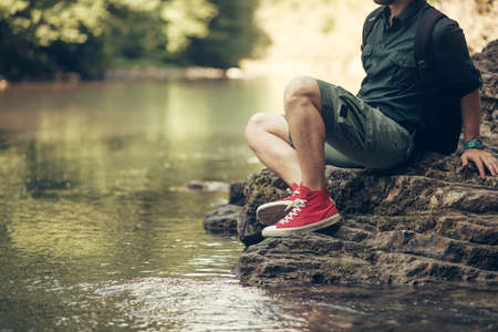 Travel concept. Unrecognizable tourist man in red sneakers, wearing travel outfit, resting on the mountain river bank in a summer wood, close upの写真素材