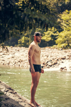 Young bearded man with a strong trained body and swimming pants stands on the rocky lake shore against the green nature. Rest tourism relaxation. Unity with nature.の写真素材