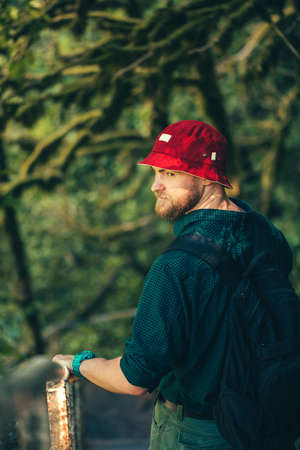 Half-size portrait of European bearded Hiker in red hat wearing backpack, over the blurred deep forest backgroundの写真素材