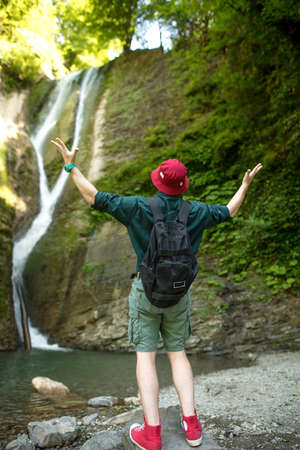European Hiker man in red hat and red sneakers with lifted hands before highland waterfall in Iceland, back view.の写真素材