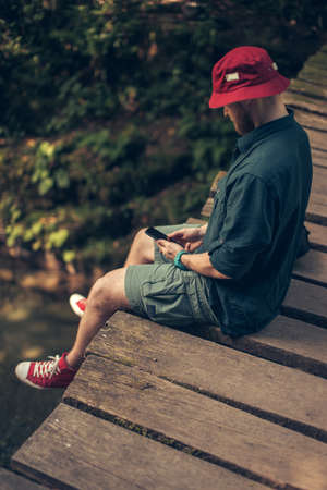 Lost adventurer man in red hat sits on old wooden bridge while using smartphone to download the map to find a way in forest. Adventure and travelling concept.の写真素材